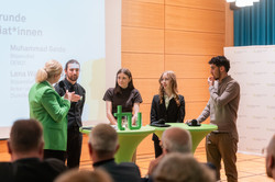 Moderatorin Britt Lorenzen leitet eine Talkrunde mit vier Stipendiatinnen und Stipendiaten. Alle stehen im Bühnenbereich an zwei Stehtischen. 