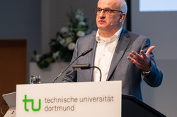 A man stands at a desk with "technische universität dortmund" written on the front.