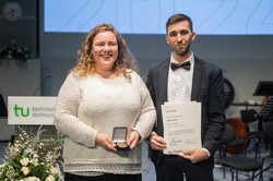 Das Bild zeigt die Asta-Vorsitzende sowie den Lehrpreis Studenten mit Medaille und Urkunde. Im Hintergrund ist ein Teil der Präsentation zu sehen sowie ein Pult mit TU-Schild und Blumengesteck vorne dran.