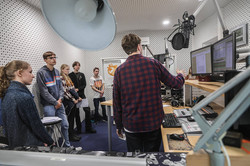 Eine Gruppe junger Leute steht in einem Radiostudio um ein Pult herum. Ein Mann steht rechts neben Monitoren und erklärt etwas.