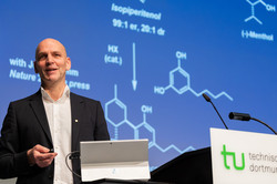 A man is standing at a podium. In front of him is a tablet, with images of molecules projected onto the wall in the background.