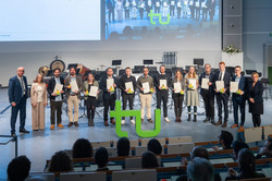 Das Bild zeigt ein Gruppenfoto der Personen mit den besten Dissertationen im Jahr 2024. In der Mitte vor den Personen steht ein großes, grünes TU-Logo. Im Vordergrund sind dunkel die Umrisse des Publikums erkennbar. Im Hintergrund sind ebenso zwei Präsentationsbilder zu sehen.