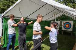 Photo: Two people practicing archery at a stand at the summer festival. Two other people explain the correct stance to them.