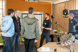 Eine Gruppe von vier Personen steht um einen Mann mit schwarzem T-Shirt, der etwas erklärt. Vorne auf dem Tisch steht eine Apparatur mit sechseckigen Spiegeln und Stäben.