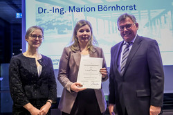 Group photo: Three people stand in front of a screen, one of them holding a certificate.