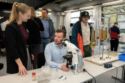 The picture shows people in front of a table with various models on it. One person is sitting in front of a microscope.