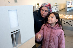 The picture shows a child in a pink jacket with a woman leaning down towards him. Both are looking at a white picture of a building. In the background is a table with a wooden model of the city.