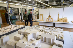 In einer Werkstatt stehen im Hintergrund Maschinen. Im Vordergrund sind Holzmodelle von Gebäuden, Gebäudeteilen und Stadtteilen ausgestellt. Zwei Männer gehen auf die Modelle zu.