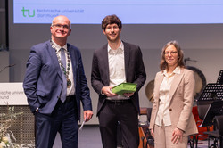 Das Bild zeigt drei Personen, die alle schick angezogen sind und in die Kamera lächeln. In der Mitte Jens Mazei der Young Academy mit einem Geschenk im grünen Papier verpackt. Im Hintergrund ist eine Präsentation zu sehen.