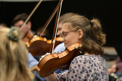 The picture shows a woman playing a violin from the side. In the background, several people can be seen in a blur, also playing string instruments.
