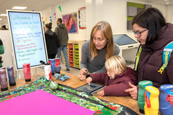 The picture shows a table with lots of utensils. On the table is a tablet being operated by a child. Two adult women are standing next to the child. On the left of the picture is a poster showing what visitors can try out at the Hyle-C stand.