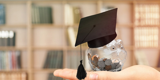 A transparent piggy bank full of coins and with a doctoral cap is held in front of a book shelf. 